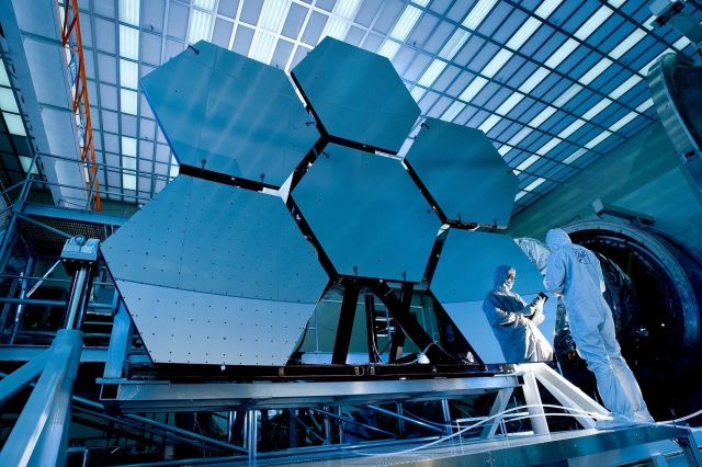 Men stand infront of a large telescope mirror and look at a tablet