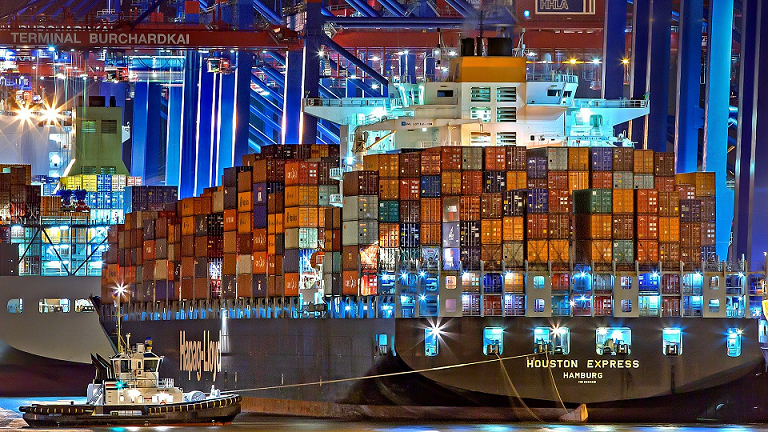 Large cargo ship in industrial port at night with a small ship in the foreground